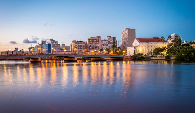 Foto panorâmica de Recife ao entardecer. Ao fundo aparecem construções antigas às margens do rio Capibaribe.